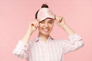 Young happy smiling female wearing striped shirt and eye mask for sleeping, keeping it on forehead and winking with one eye, isolated on pink background
