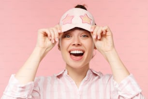 Young excited laughing female holding sleeping mask on forehead and looking from under it, having fun on pyjama party, isolated on pink background