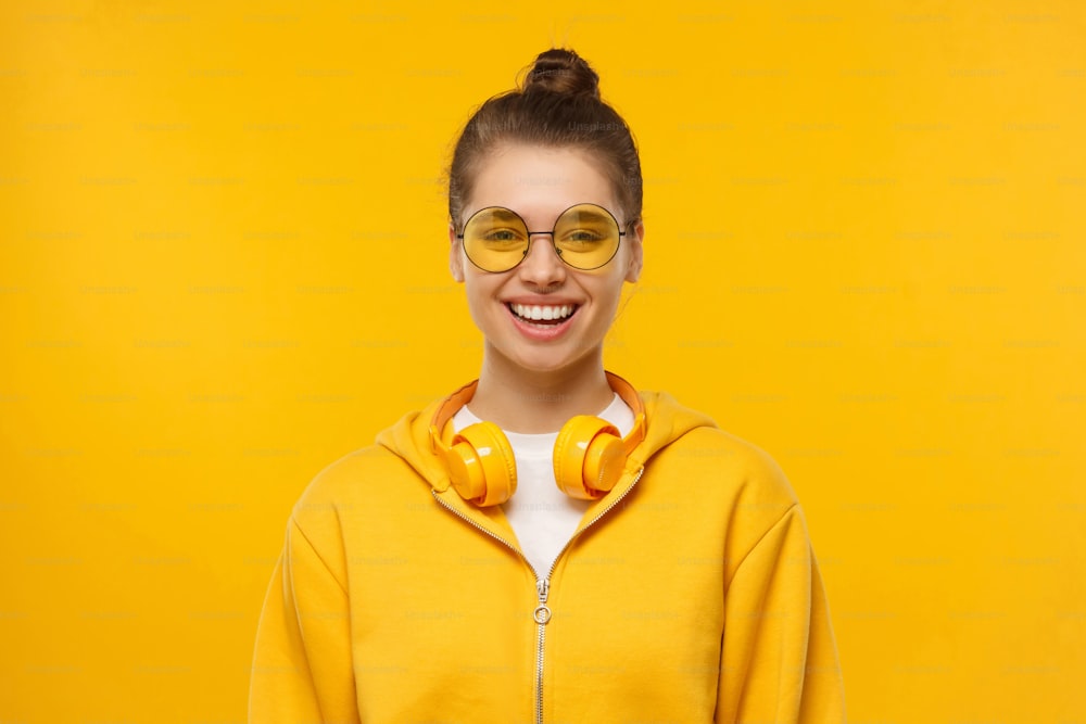 Junges lachendes Teenie-Mädchen mit Kapuzenpullover, runder farbiger Brille und kabellosen Kopfhörern um den Hals, isoliert auf gelbem Hintergrund