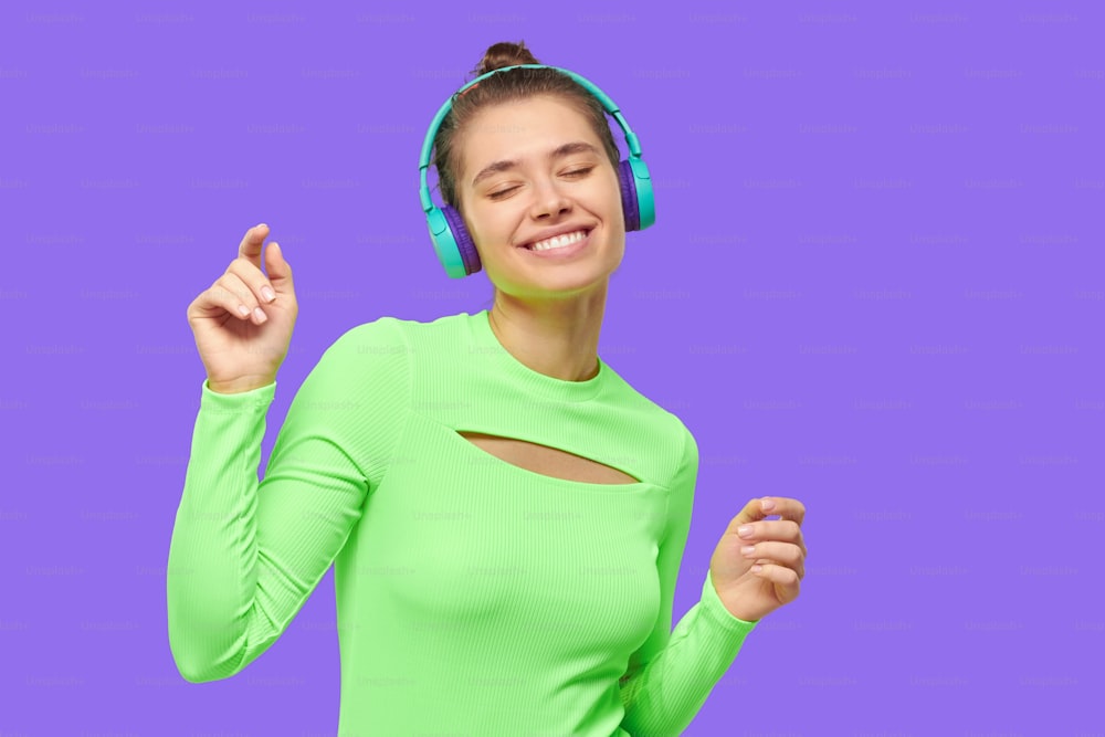 Young happy teen girl in green neon top, wearing wireless headphones and dancing to music with eyes closed, isolated on purple background
