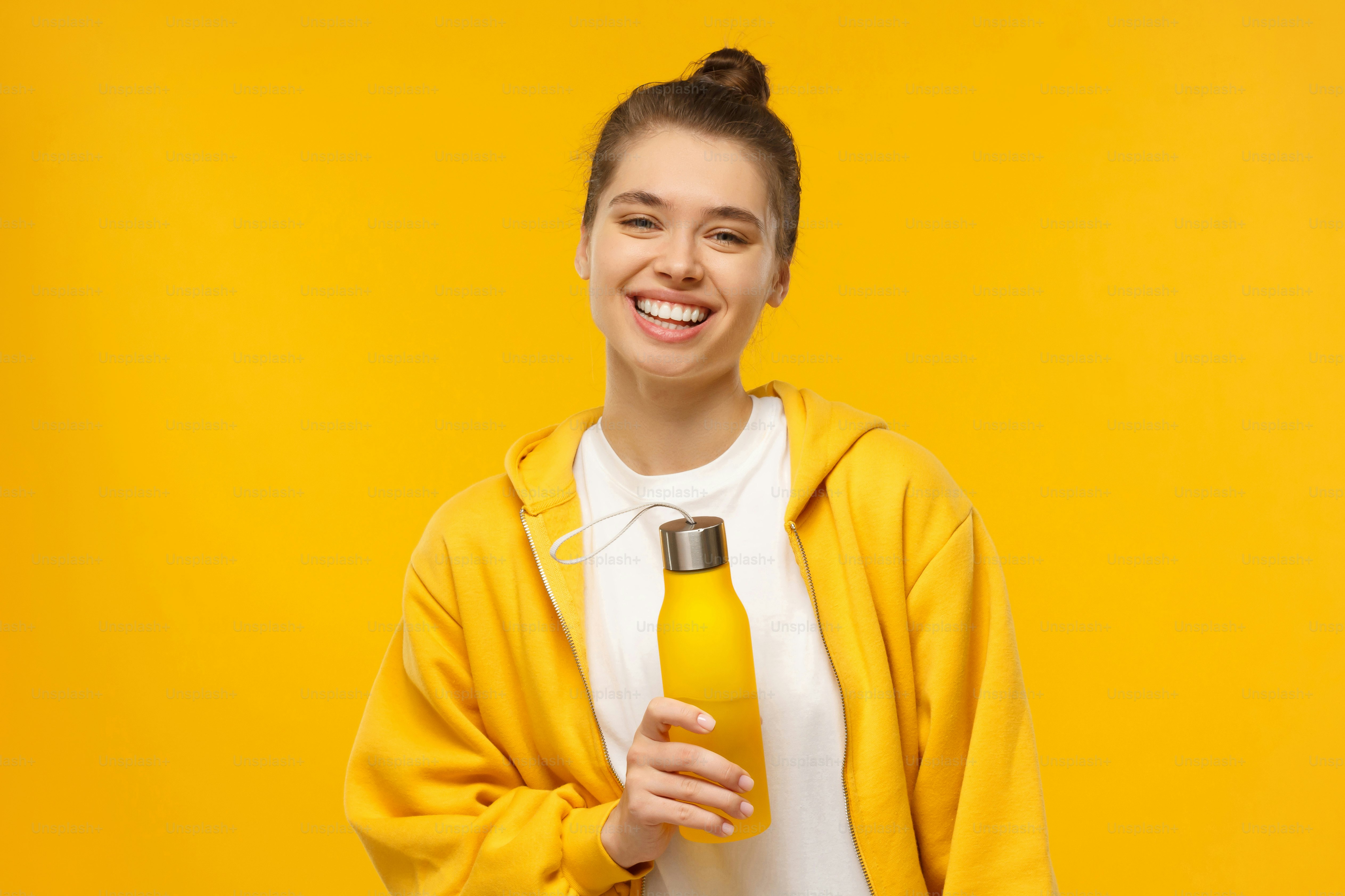 Desportista jovem no capuz brilhante e t-shirt branca, segurando garrafa de água ecológica reutilizável, isolada no fundo amarelo