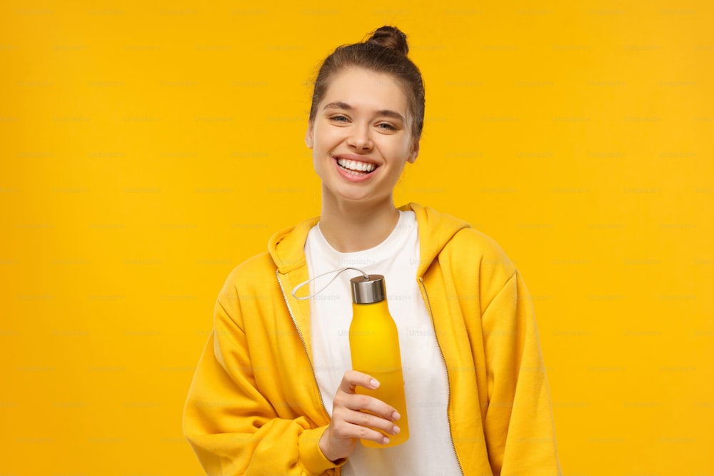 Young sportswoman in bright hoodie and white t-shirt, holding reusable eco water bottle, isolated on yellow background