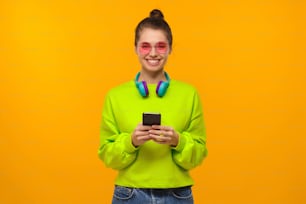 Ritratto di ragazza adolescente che indossa felpa verde neon, occhiali rosa e cuffie sul collo, in piedi con il telefono in mano, isolato su sfondo giallo