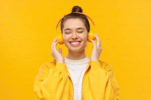Joven niña feliz sonriendo y riendo con los ojos cerrados, disfrutando de la música en auriculares inalámbricos, aislada sobre fondo amarillo