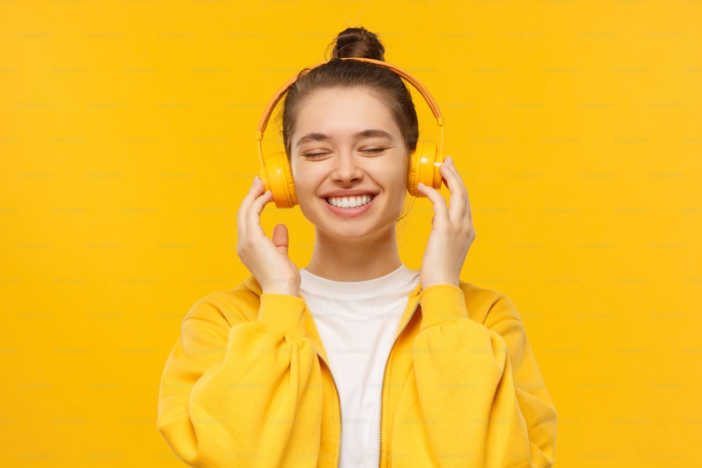 Young happy girl in smiling and laughing with closed eyes, enjoying music in wireless headphones, isolated on yellow background