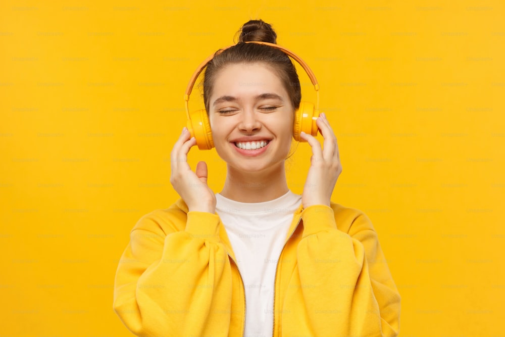 Young happy girl in smiling and laughing with closed eyes, enjoying music in wireless headphones, isolated on yellow background