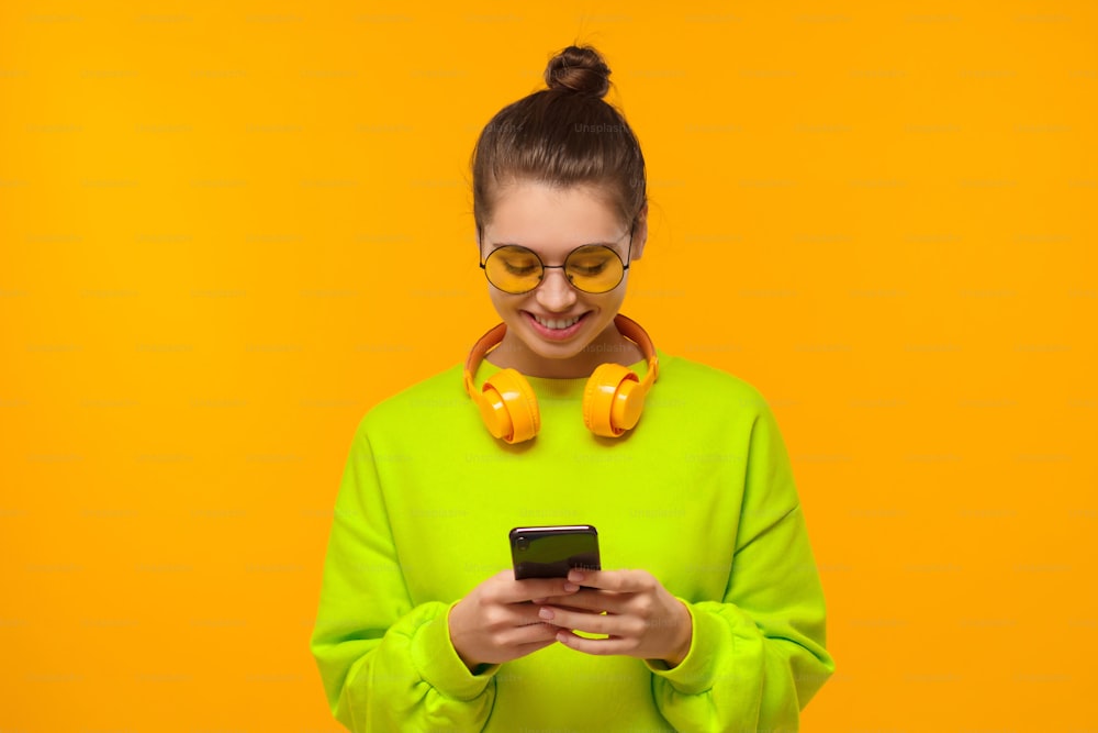 Junge Frau mit Kopfhörern um den Hals, schaut mit einem Lächeln auf den Bildschirm des Smartphones, isoliert auf gelbem Hintergrund