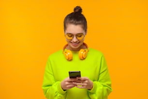 黄色い背景に笑顔でスマートフォンの画面を見る首にヘッドフォンを持つ若い女性