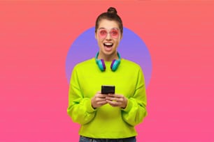 Giovane donna che indossa occhiali rosa, felpa verde neon e cuffie intorno al collo, eccitata dal contenuto sullo schermo del telefono