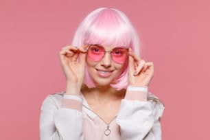 스웨트셔츠, 가발, 안경을 쓰고 파티를 즐기고 분홍색 배경에 고립된 멋진 모습을 하고 웃고 있는 젊은 트렌디한 소녀의 클로즈업 초상화