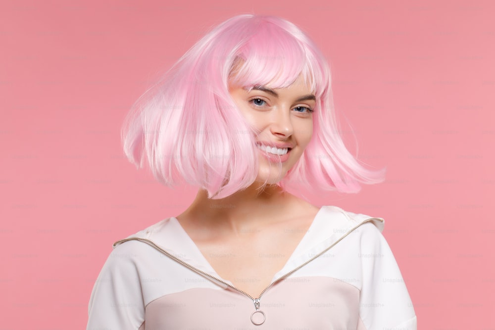 Portrait of young happy teenage girl wearing sweatshirt and wig, moving her head so hair is flying to sides, feeling cheerful, isolated on pink background