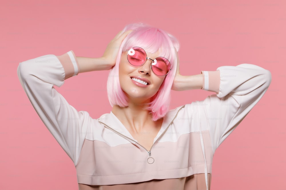 Young happy girl in sweatshirt, wig and eyeglasses, holding head, chilling with closed eyes, dancing to music at party, isolated on pink background
