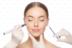 Mujer joven lista para procedimientos de belleza con los ojos cerrados. Los médicos sostienen el marcador de la piel y la jeringa de inyección, aislados sobre fondo blanco
