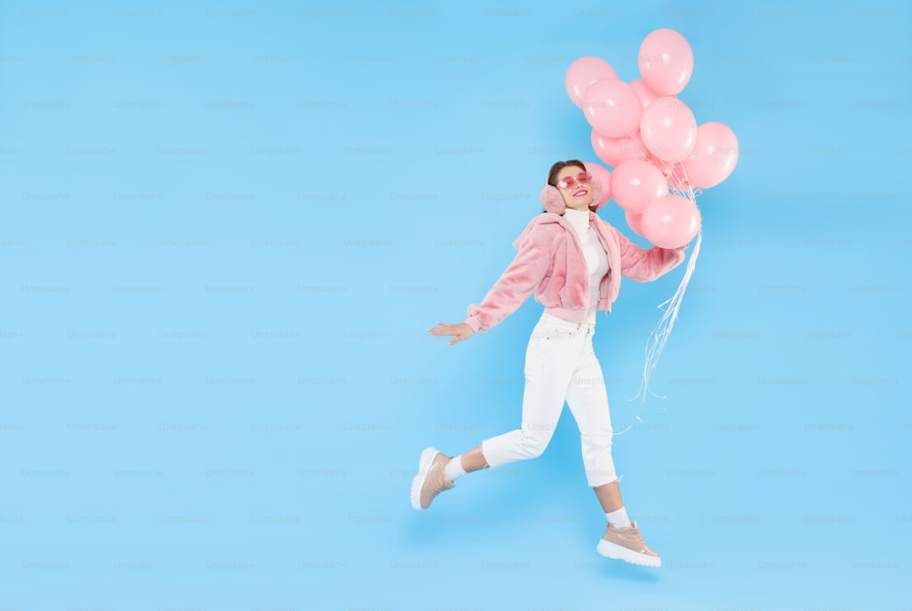 Mujer joven feliz con abrigo rosa esponjoso, gafas de colores y orejeras, corriendo y saltando con globos en la mano, aislada sobre fondo azul