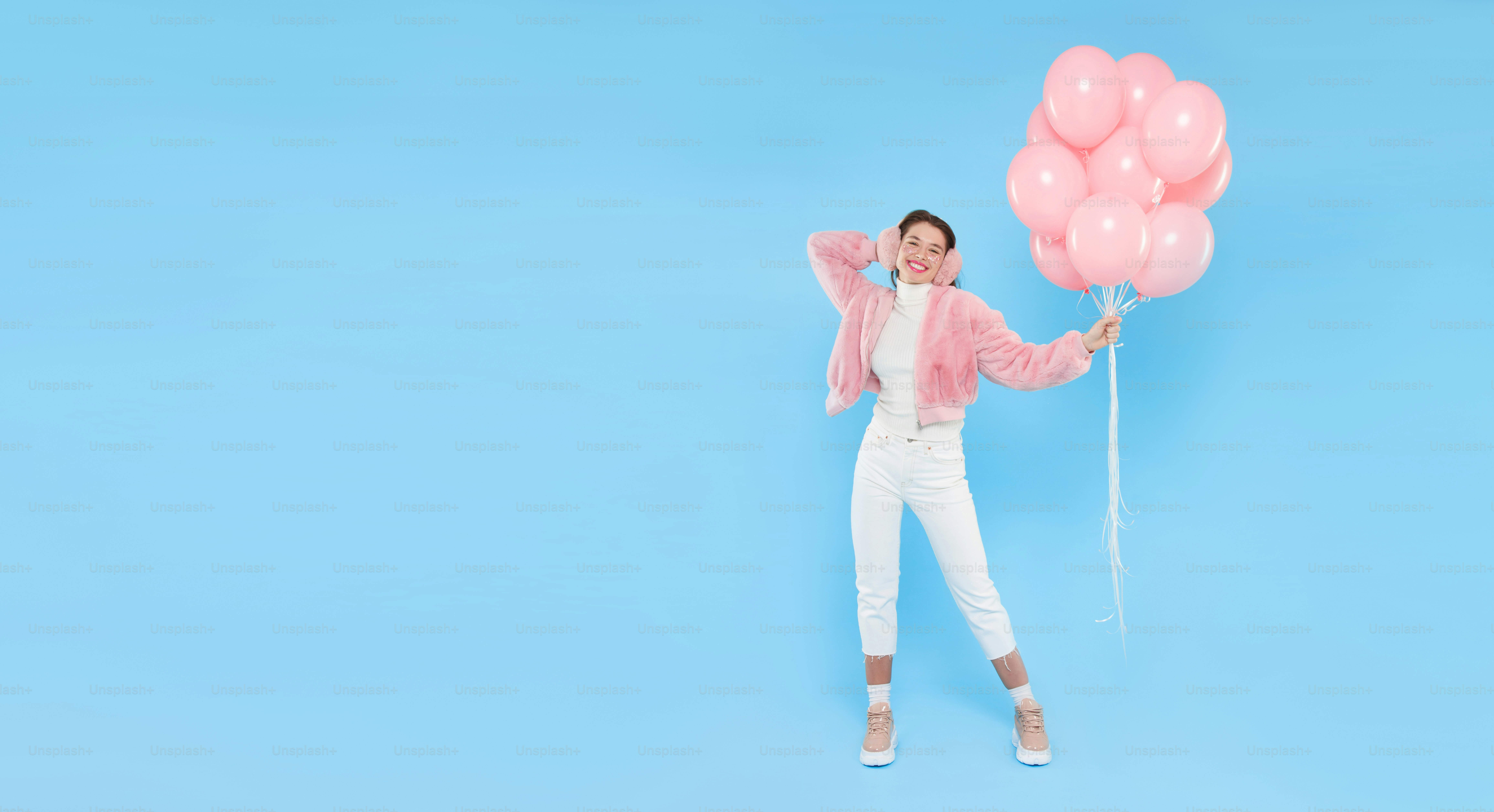 Horizontal banner of young happy girl wearing pink furry coat and earmuffs, standing with pink birthday balloons, isolated on blue background, copy space on left