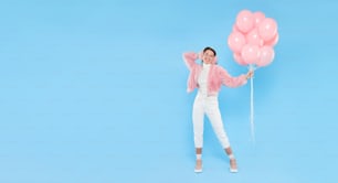 Horizontal banner of young happy girl wearing pink furry coat and earmuffs, standing with pink birthday balloons, isolated on blue background, copy space on left