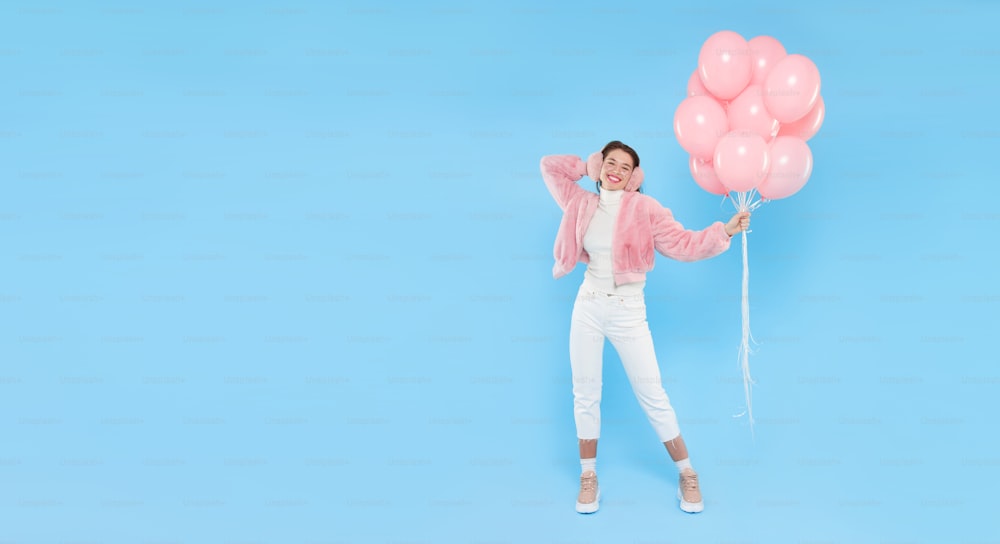 Estandarte horizontal de una joven feliz con abrigo peludo rosa y orejeras, de pie con globos de cumpleaños rosas, aislado sobre fondo azul, espacio de copia a la izquierda