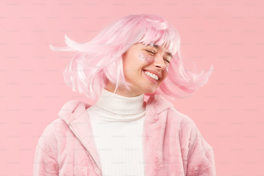 Junges glückliches Mädchen mit geschlossenen Augen im Pelzmantel, tanzt und bewegt den Kopf, so dass ihr Haar fliegt, isoliert auf rosa Hintergrund