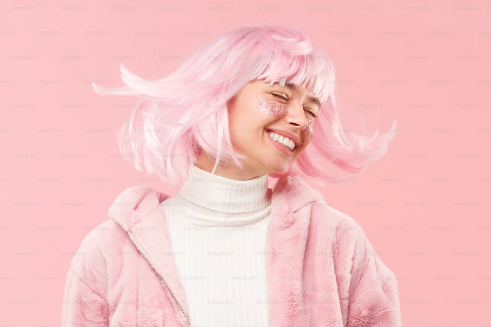 Young happy girl with closed eyes in fur coat, dancing and moving head so her hair is flying, isolated on pink background