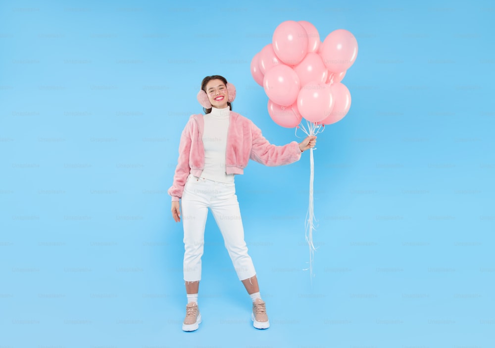 Young positive girl wearing pink fluffy winter bomber and ear warmers, standing with bunch of pink balloons, celebrating her birthday, isolated on blue background