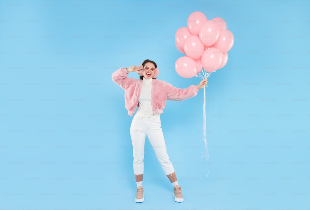 Ganzkörperporträt einer jungen glücklichen Frau, die rosa flauschige Bomberjacke und Ohrwärmer trägt und mit rosa Geburtstagsballons steht, isoliert auf blauem Hintergrund