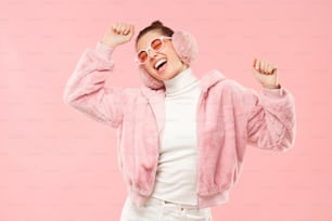 Giovane ragazza eccitata che indossa un caldo cappotto peloso, paraorecchie e occhiali colorati, muovendo le braccia a suoni di musica, isolata su sfondo rosa