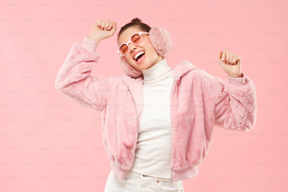 Junges, aufgeregtes Mädchen in warmem Pelzmantel, Ohrenschützern und farbigen Brillen, bewegt die Arme zu Klängen der Musik, isoliert auf rosa Hintergrund