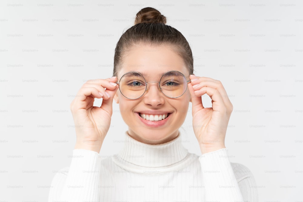 Young woman holding glasses that she is wearing, smiling, looking straight at camera, feeling relaxed and satisfied with good eyesight, isolated on gray background