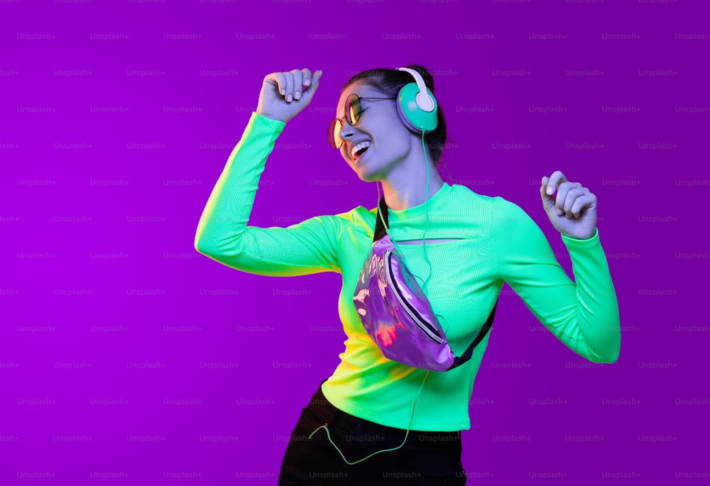 Young girl dancing to sounds of music listening in headphones, dressed in neon green top, isolated on purple background
