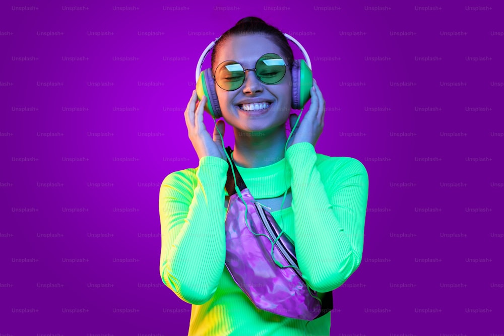 Retrato de una joven feliz con top verde neón, con auriculares, escuchando su canción favorita, aislada sobre fondo púrpura