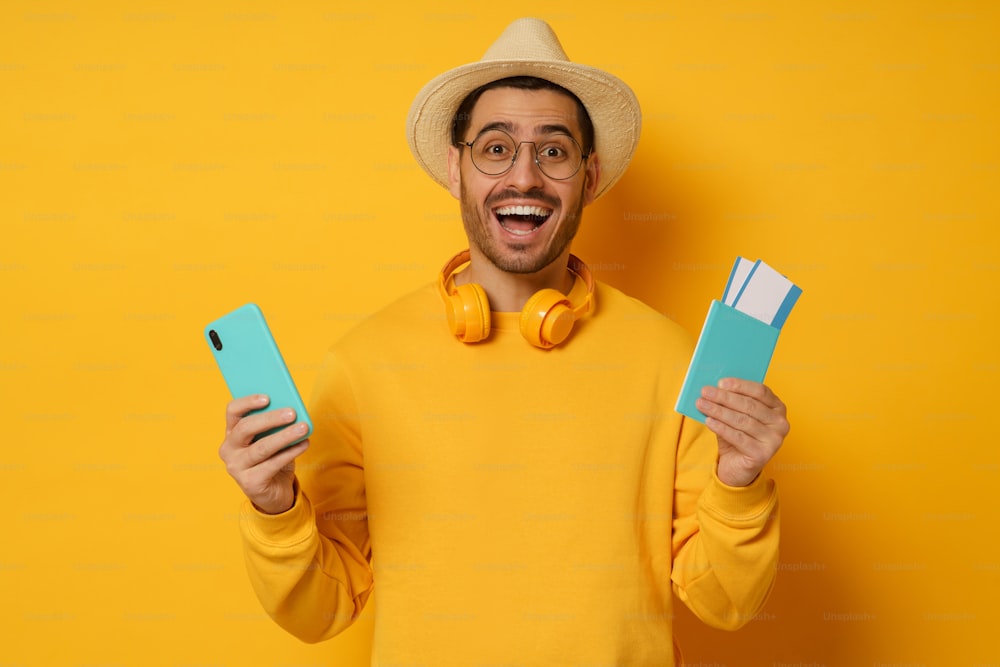 Young excited man student tourist holding passport and smart phone with travel app for buying tickets online, isolated on yellow background