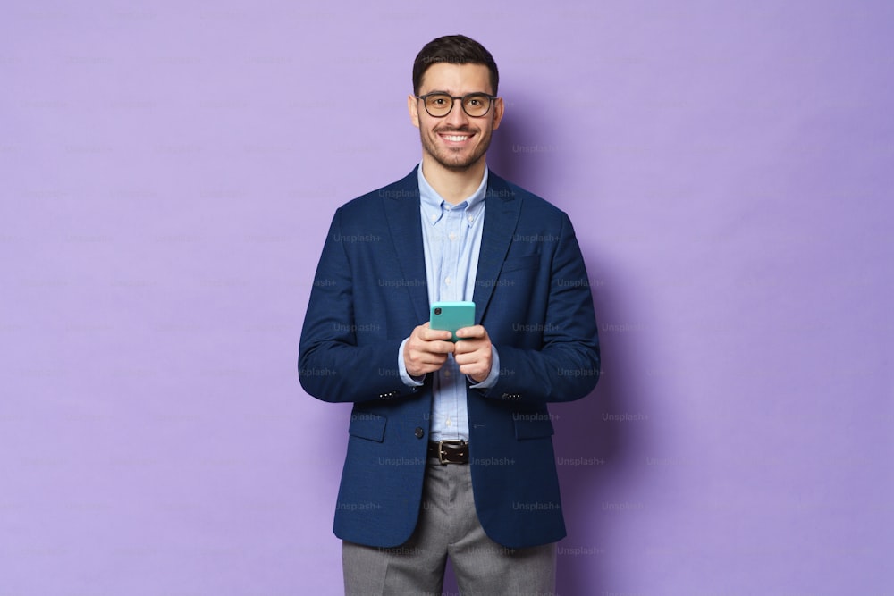 Business man looking at camera with happy face, holding cellphone in hands, communicating with colleagues and partners, smiling, isolated on purple background