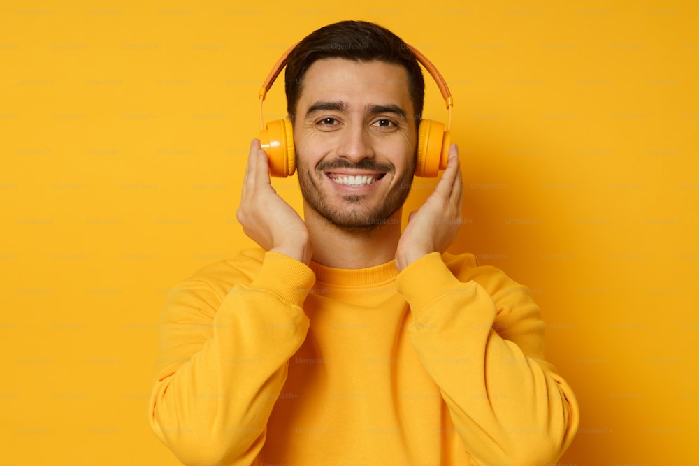Junger Mann, der Musik hört, drahtlose Kopfhörer und ein helles Sweatshirt trägt und Lieblingstitel genießt, isoliert auf gelbem Hintergrund