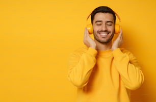 Handsome man relaxing to sounds of favorite music tracks via wireless headphones, dressed in sweatshirt, keeping eyes closed, isolated on yellow background