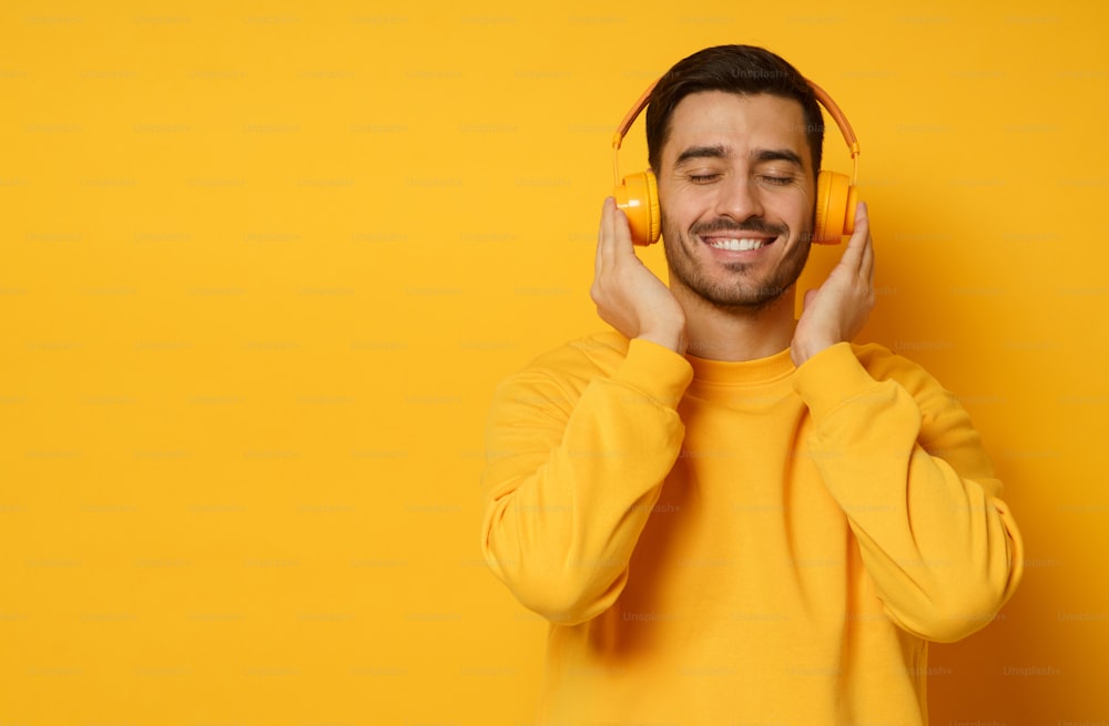잘 생긴 남자는 무선 헤드폰을 통해 좋아하는 음악 트랙의 소리에 휴식을 취하고, 스웨트 셔츠를 입고, 눈을 감고, 노란색 배경에 고립되어 있습니다