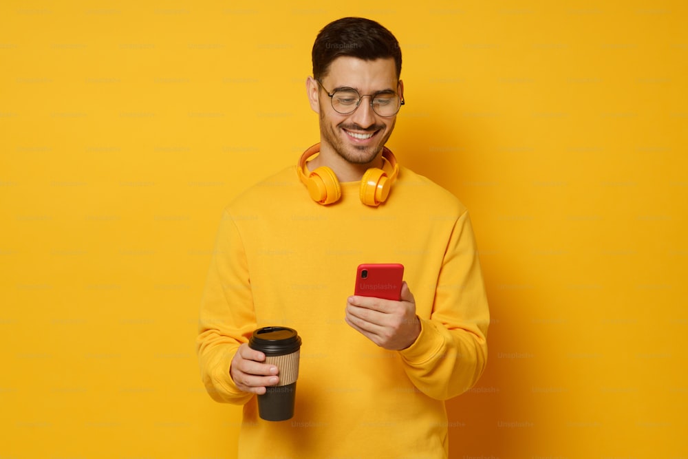 Millennial man in glasses, enjoying messages from friends via phone screen, smiling happily, drinking coffee, headphones round neck, isolated on yellow background