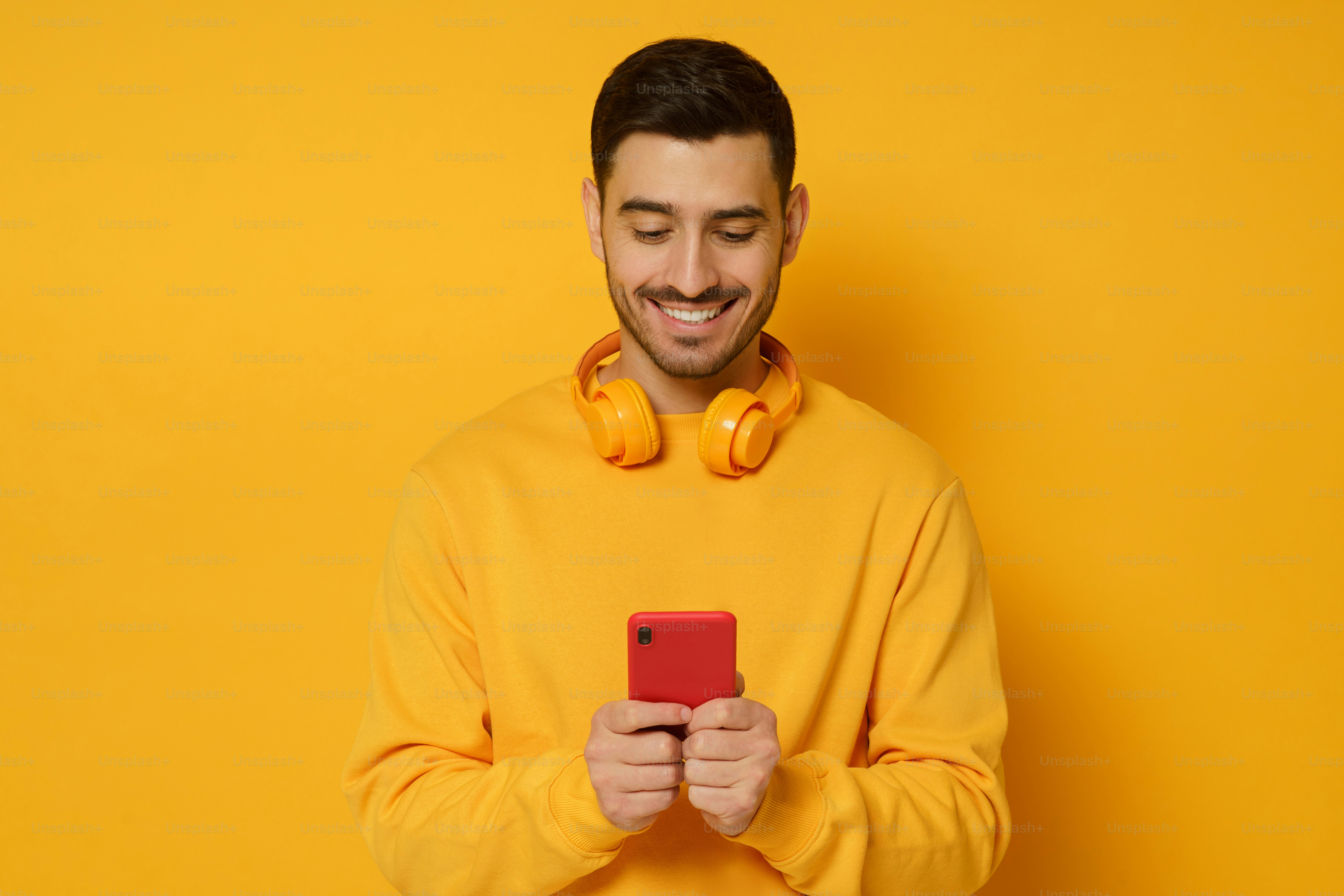 Young trendy man wearing sweatshirt and wireless headphones, smiling while looking at content on phone screen, isolated on yellow background
