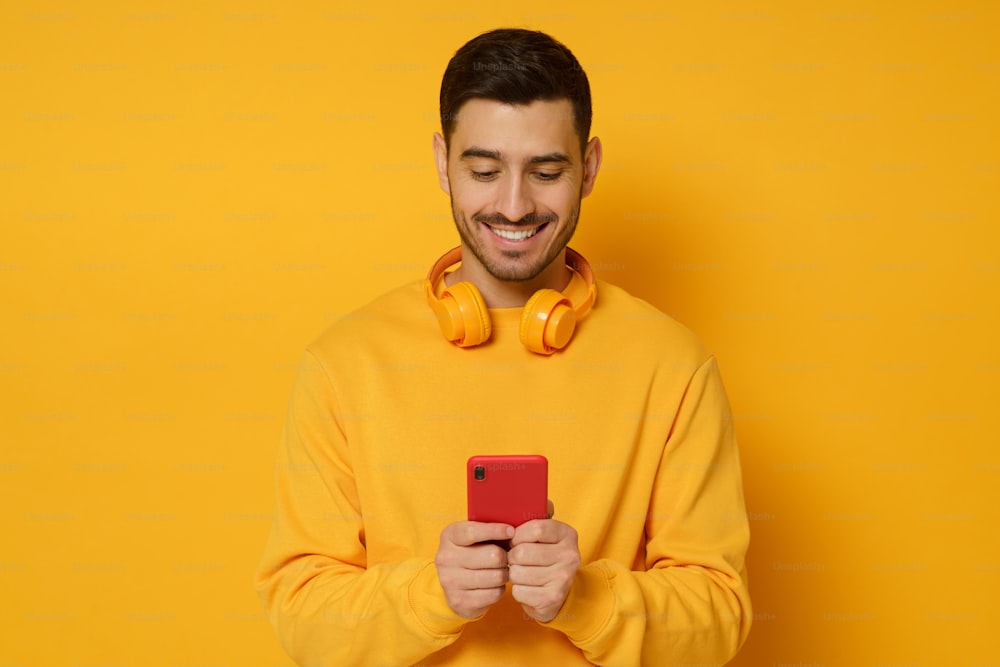Joven hombre moderno con sudadera y auriculares inalámbricos, sonriendo mientras mira el contenido en la pantalla del teléfono, aislado sobre fondo amarillo