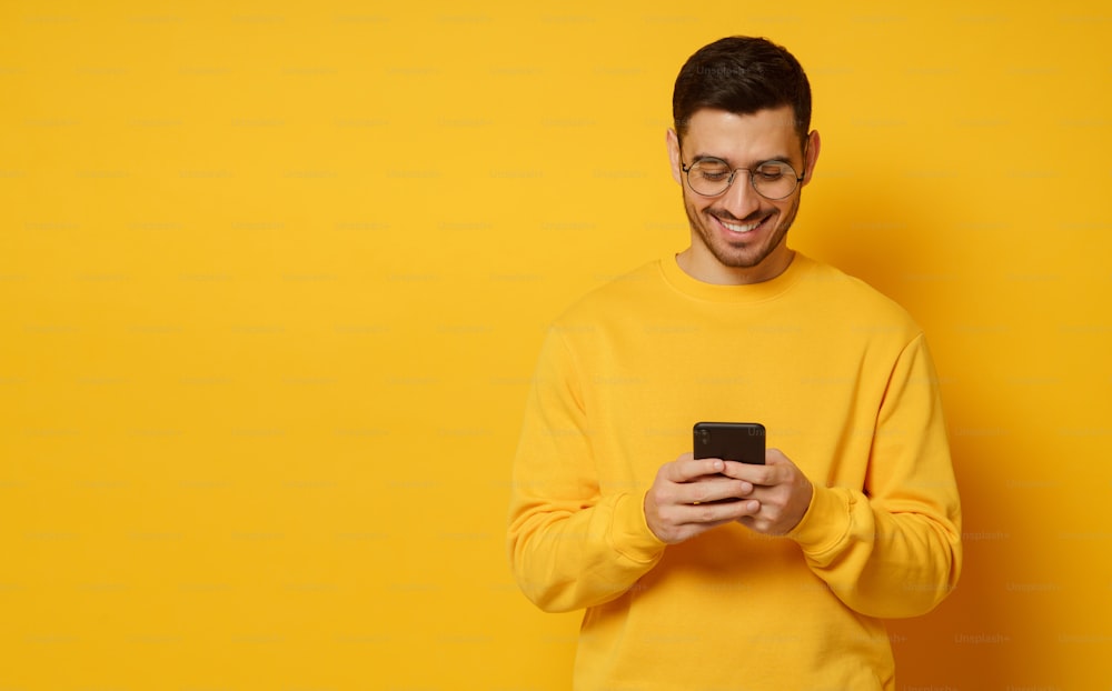 Horizontal banner of young man in bright sweatshirt, holding smartphone in both hands, laughing at content on screen, isolated on yellow background, copy space on left
