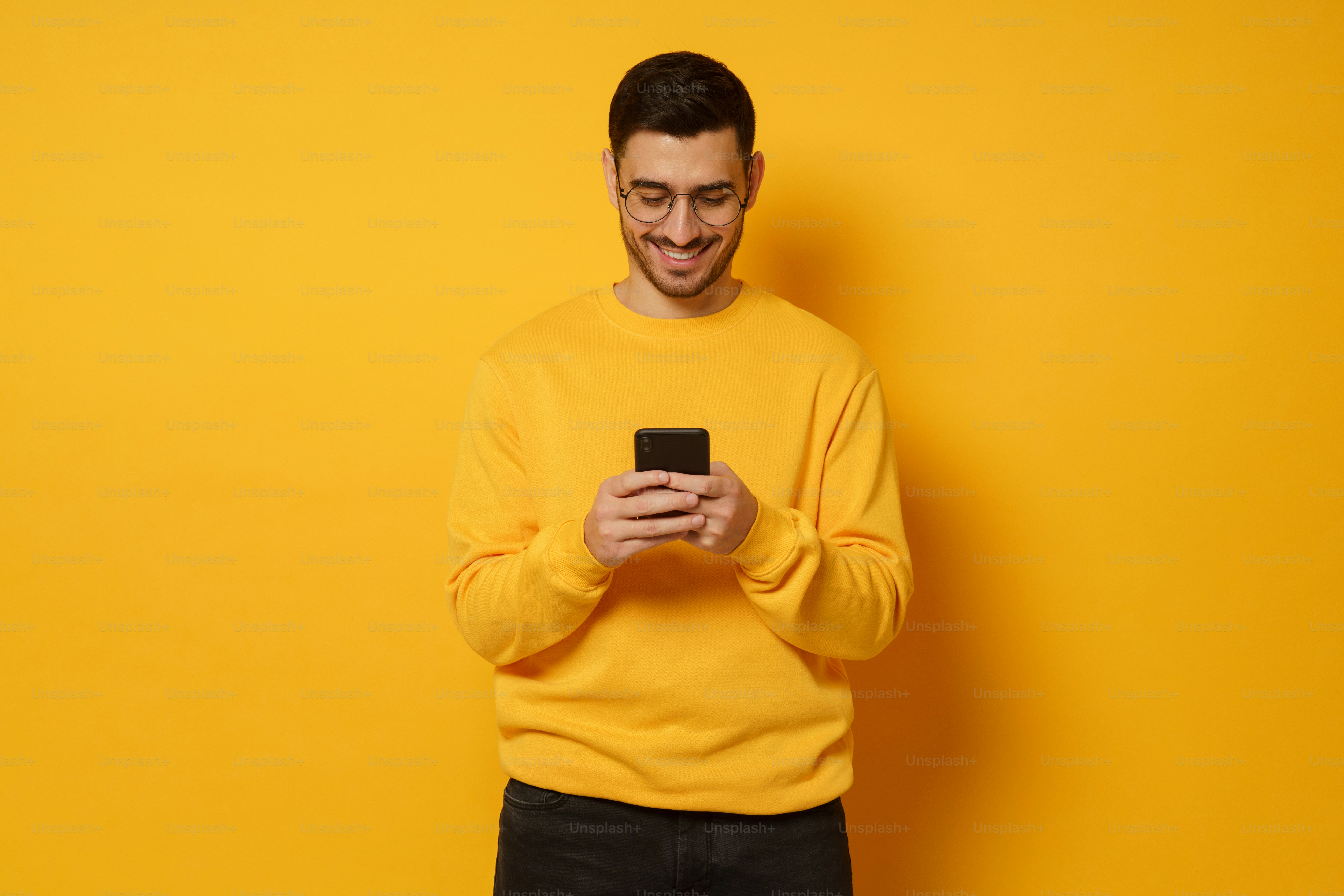 黄色い背景に明るいスウェットシャツを着て、スマートフォンで友達とテキストメッセージを交換する若いハンサムな男性
