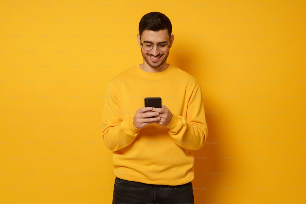 Young handsome man exchanging text messages with friends via smart phone, having fun, wearing bright sweatshirt, isolated on yellow background