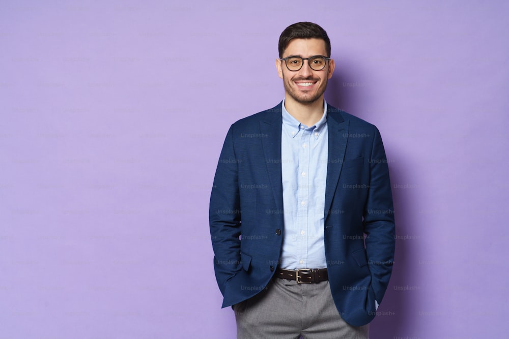 Jovem empresário vestindo roupas formais e óculos, de pé isolado contra o fundo roxo em pose descontraída, sorrindo amigável, espaço de cópia à esquerda