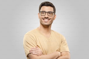 Photo of guy in beige T-shirt and glasses, looking aside with dreamful expression and smile, standing with arms crossed, isolated on gray background