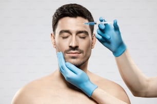 Hombre joven recibiendo una inyección para el cuidado de la piel en la frente, sintiéndose tranquilo, confiando en el médico en la clínica, con los ojos cerrados, aislado sobre fondo gris. Lifting facial hombre