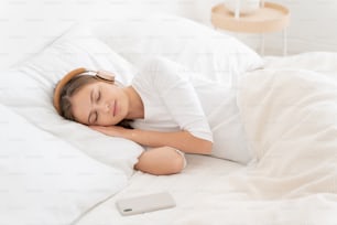 Relaxed lady dreaming on white bed, wearing white top and wireless headphones, listening to audiobook on smartphone that made her sleep