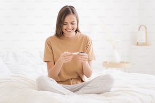 Mujer joven sentada en la cama a la luz del día, mirando la prueba de embarazo que muestra un resultado positivo, sonriendo felizmente sobre la idea del niño