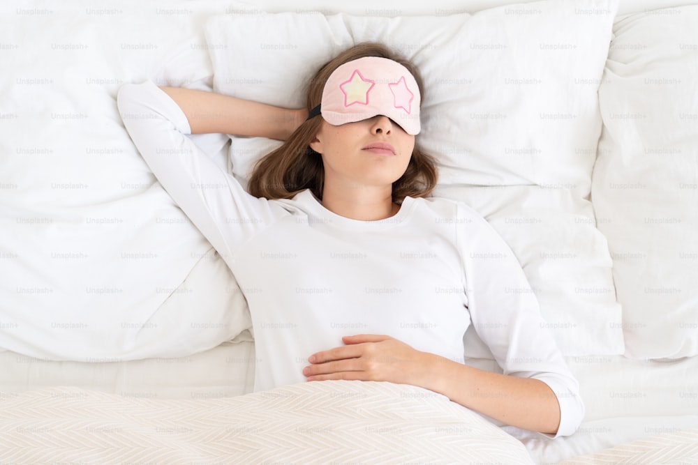 Young female dreaming in white bed under blanket in morning, wearing sleeping mask that covers her eyes fully, relaxing in free time