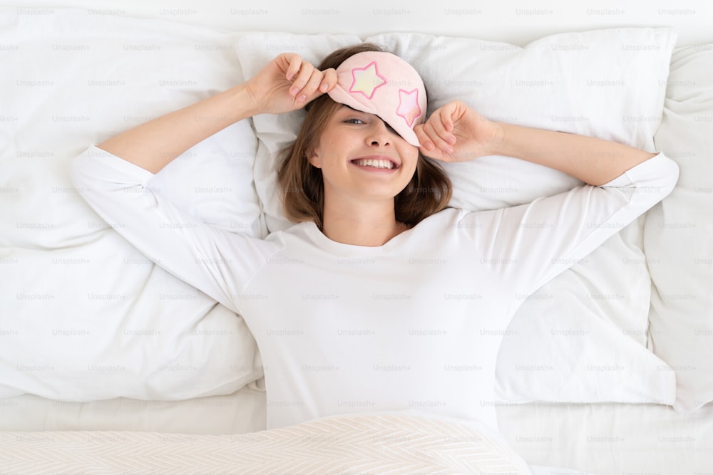 Young woman removing sleeping mask from eyes after night sleep, lying in bed in morning, looking at camera with happy face, aspiring for great positive day