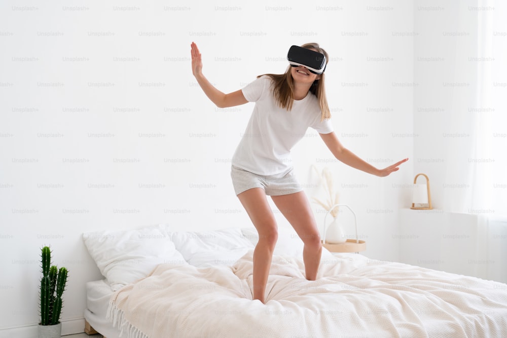 Mädchen, das die Möglichkeiten der virtuellen Realität erkundet, morgens im Bett steht, Pyjama und VR-Headset trägt, sich in der Spielumgebung bewegt und viel Spaß hat