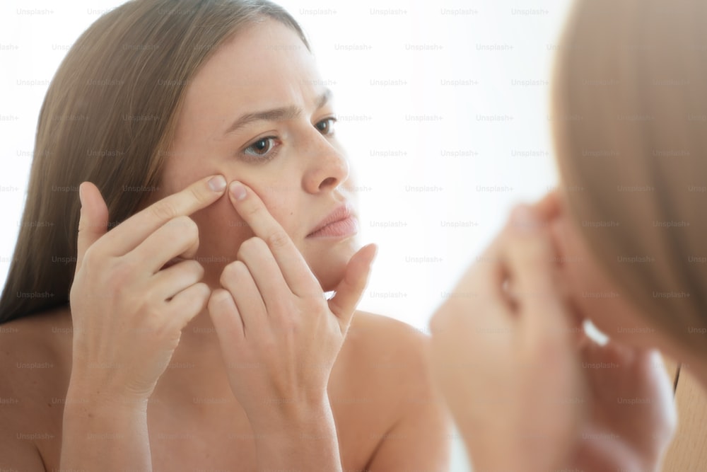 Young female unsatisfied with state of her skin, squeezing pimple in front of mirror, trying to cope with facial problems before important meeting or date